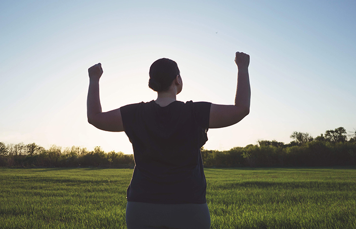 Woman in a black T-shirt is standing on a green meadow having her hands up in the air; clear blue sky, sun is rising