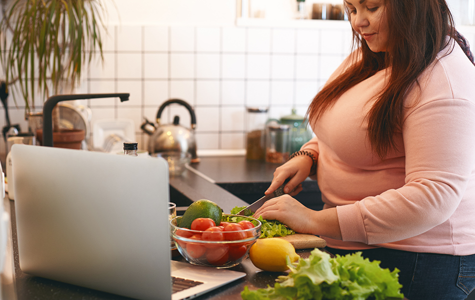 Overweight woman using laptop to watch video recipe while making vegan vitamin avocado salad, slicing leaf lettuce on wooden cutting board. Healthy food, weight loss, dieting and nutrition concept