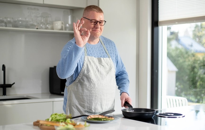 Mies paistaa hellan ääressä lohta ja näyttää OK-käsimerkkiä