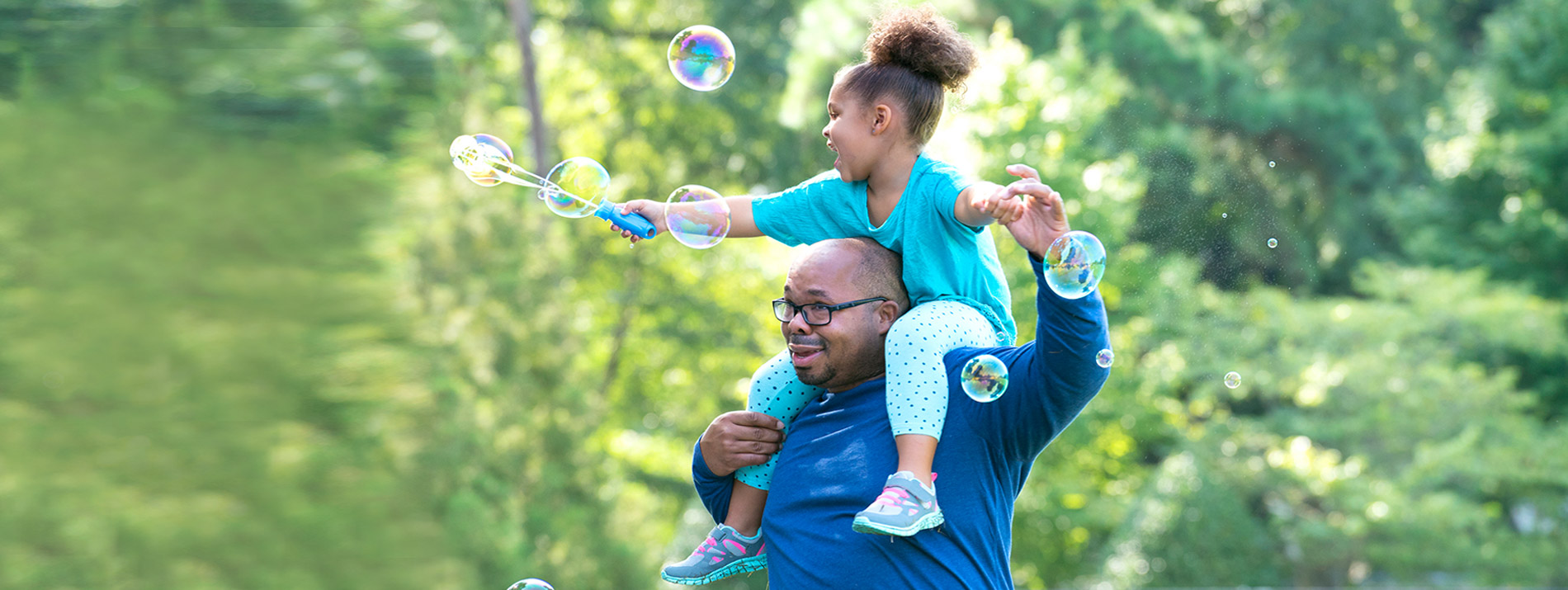 A dad playing with his daughter that is sitting on his shoulders playing with bubbles.