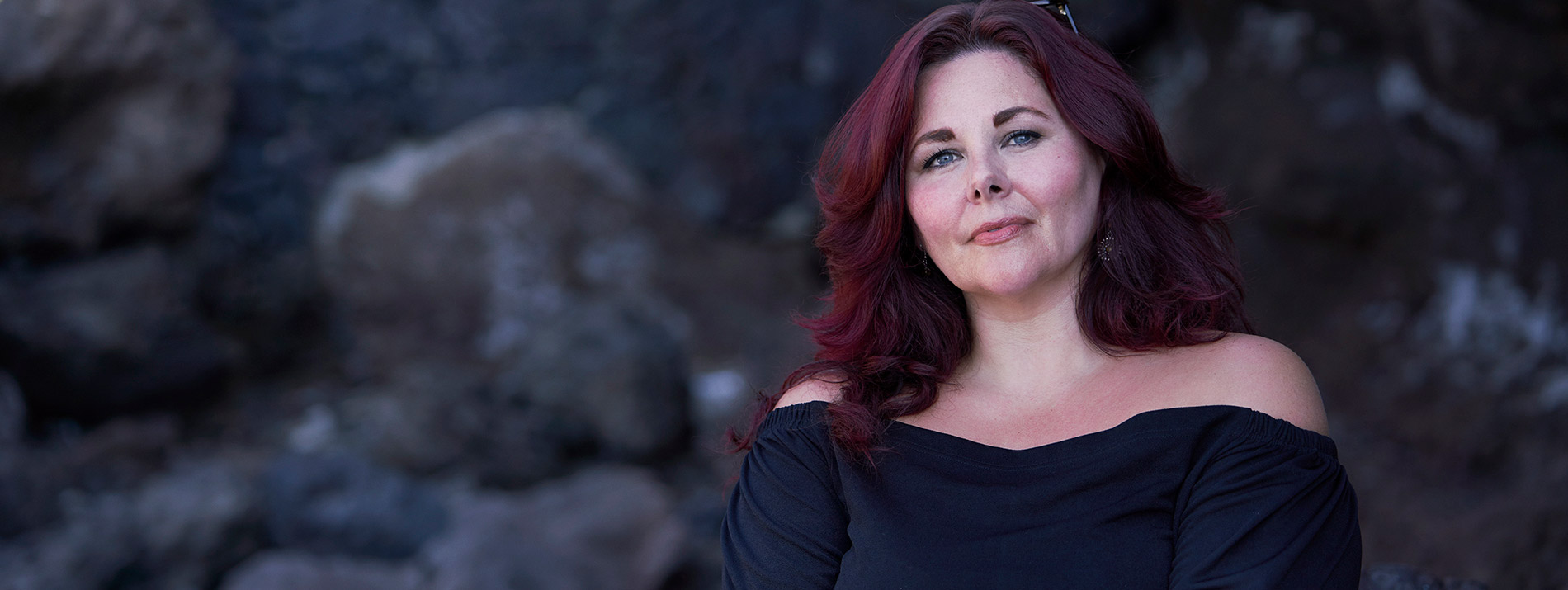 Right-aligned portrait of a confident looking woman in a black dress with a rock background.