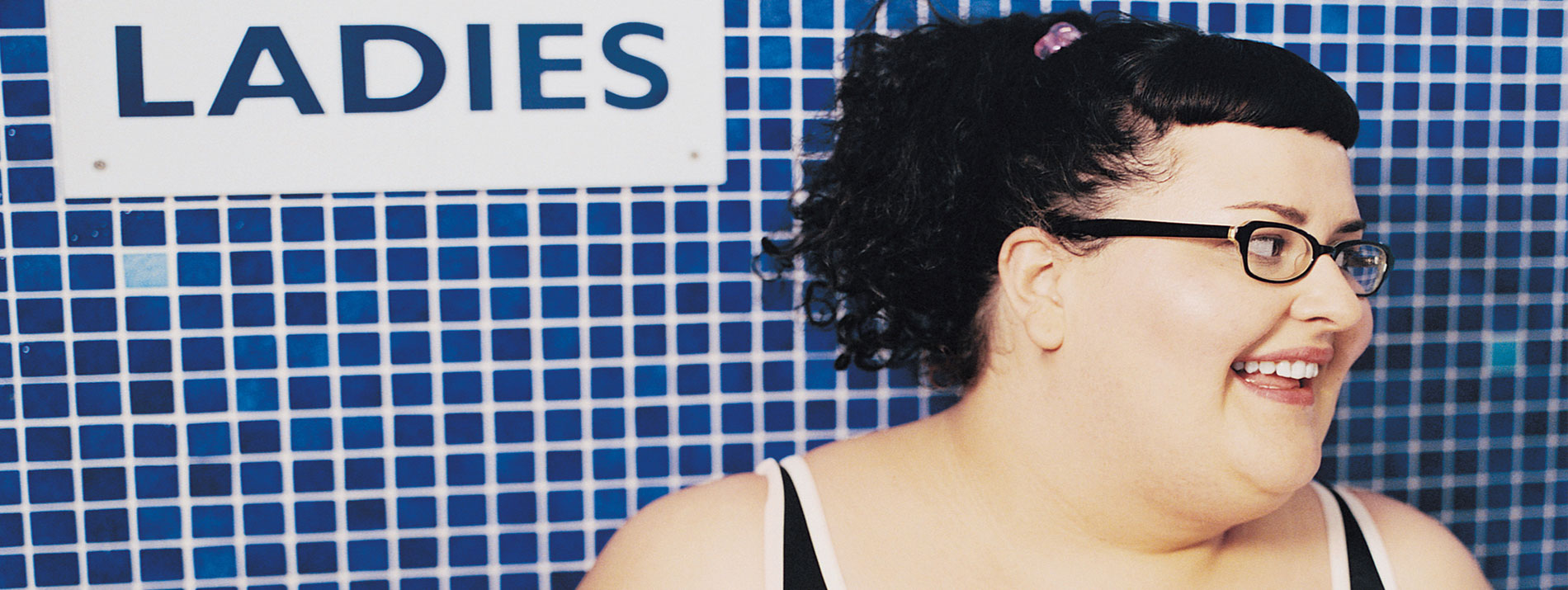 A smiling woman standing in front of a tiled wall with a 'Ladies' sign hanging on it.