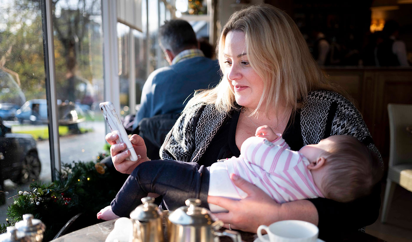 Sarah in the cafe with baby in her arms and looking at her mobile phone