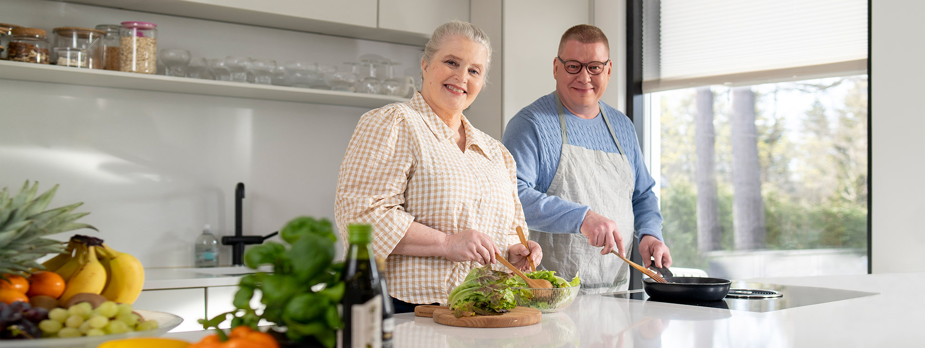 Nainen ja mies valmistavat keittiössään ruokaa, työtasolla on tuoretta salaattia ja hedelmiä